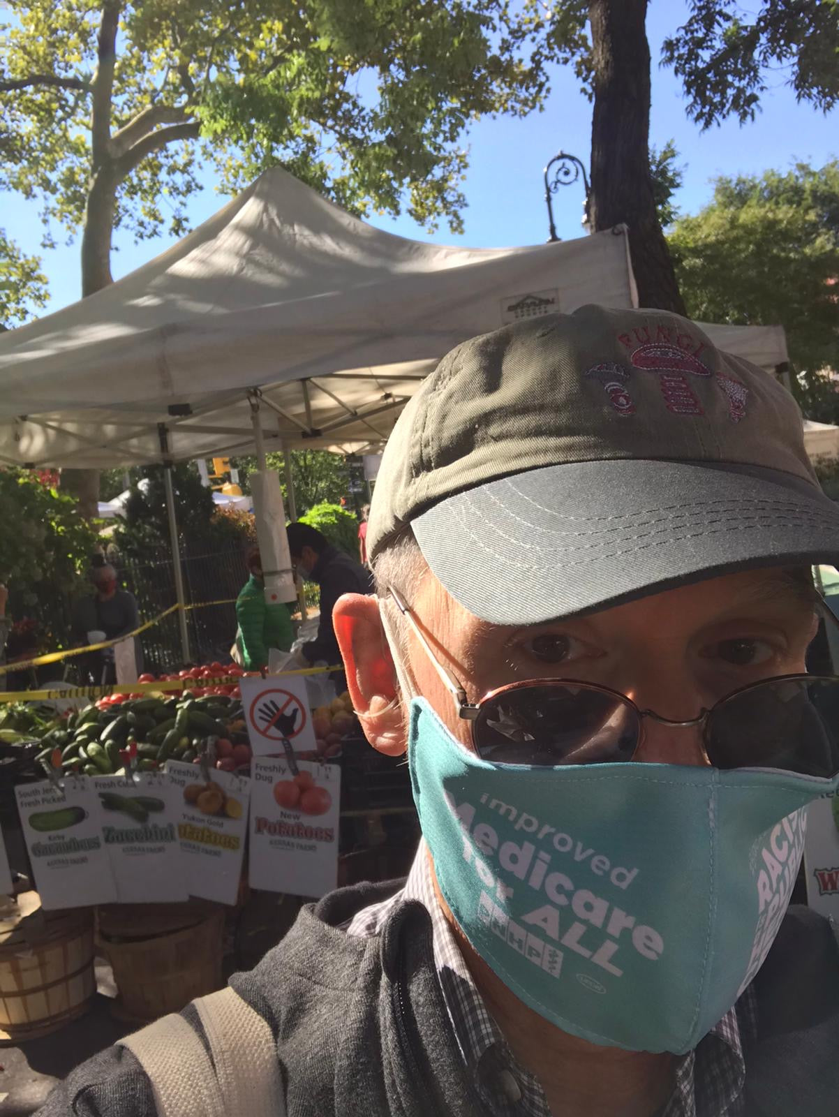 Photo of white-presenting man wearing a canvas green style baseball cap. He has sunglasses pulled down his nose - we can see his eyes. He appears to be at a farmer's market - there is a white farmstand-like tent behind him with vegetables. He is wearing a light teal face mask that says improved Medicare for ALL.