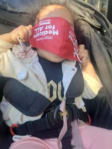 Photo of a baby in a stroller holding the red NY Health Act x improved Medicare for All face mask upside down over it's face