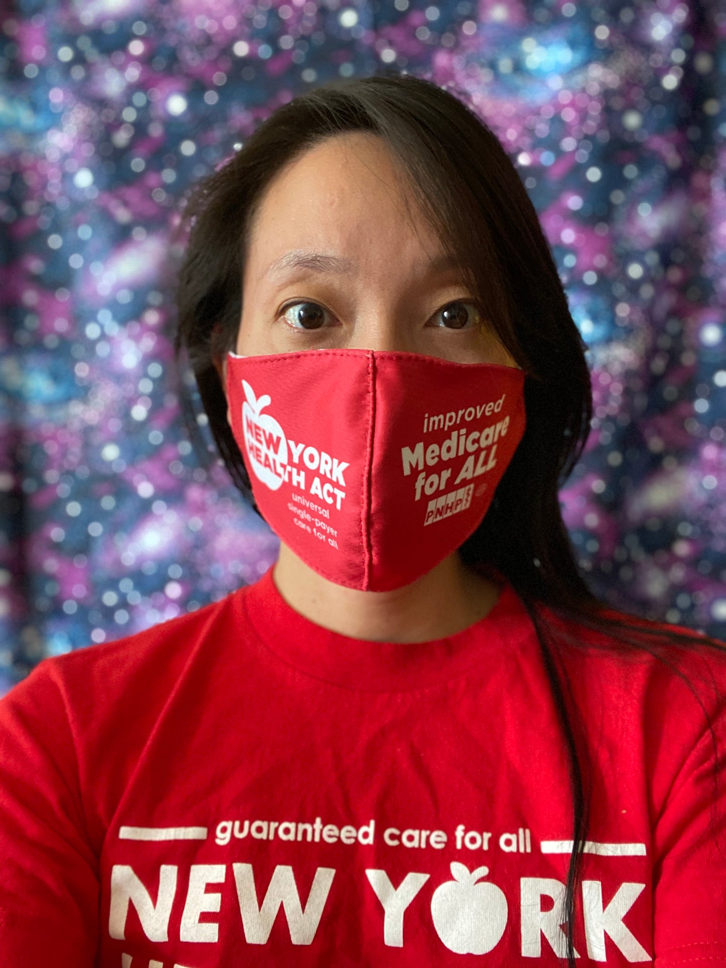 Photo of East Asian presenting  woman with long black hair and standing in front of a purple/blue/white speckled background. They are wearing a red NY Health Act face mask. The left side says "New York Health Act universal single-payer care for all" in white letters. The right side says "improved Medicare for All" in white letters and has the PNHP logo and union bug at bottom. 