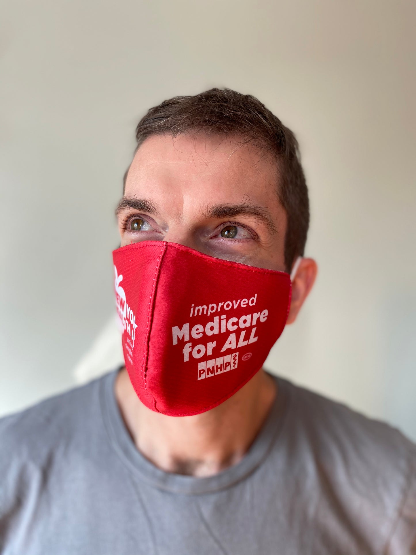 Photo of a white-presenting man looking towards the upper left, wearing a gray tshirt, has light brown hair. He is wearing the red NY Health Act face mask, showing mostly the right side of the face mask that says "improved Medicare for All"
