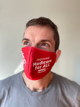 Load image into Gallery viewer, Photo of a white-presenting man looking towards the upper left, wearing a gray tshirt, has light brown hair. He is wearing the red NY Health Act face mask, showing mostly the right side of the face mask that says &quot;improved Medicare for All&quot;
