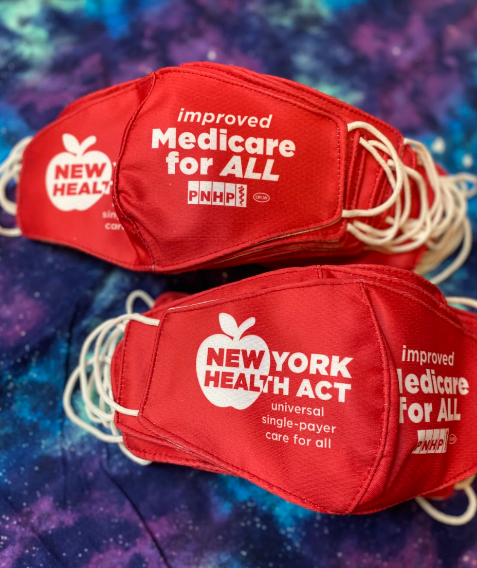 Photo of two piles of the red NY Health Act face masks laid on top of fabric that is galaxy-patterned, purple, blue, white colors. The left side of the red NY Health Act face mask says "New York Health Act" "universal single-payer care for all." The right side of the red NY Health Act face mask says "improved Medicare for All" and has the PNHP logo and union bug - all inn white.
