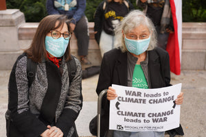 Photo of two women sitting outside. One woman has shoulder length brown hair and is wearing brown tortoise patterned glasses. The other has chin length white hair and is wearing thin silver rimmed oval  glasses. Both are white-presenting and appear to be smiling under their masks. Both are wearing the light teal improved Medicare for All face mask. Woman with white  hair is holding a sign that says "WAR leads to CLIMATE CHANGE.  CLIMATE CHANGE leads to WAR"