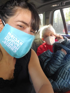 Photo of two women sitting inside a car. Woman on left has dark brown/black hair pulled back with some short bangs. The other woman has white short hair. Both women are white-presenting and appear to be smiling. Woman on left is wearing the light teal improved Medicare for all face mask, showing the side that says #BlackLivesMatter Racism Is A Public Health Crisis #MedicareForAll. The other woman is wearing a white face mask.
